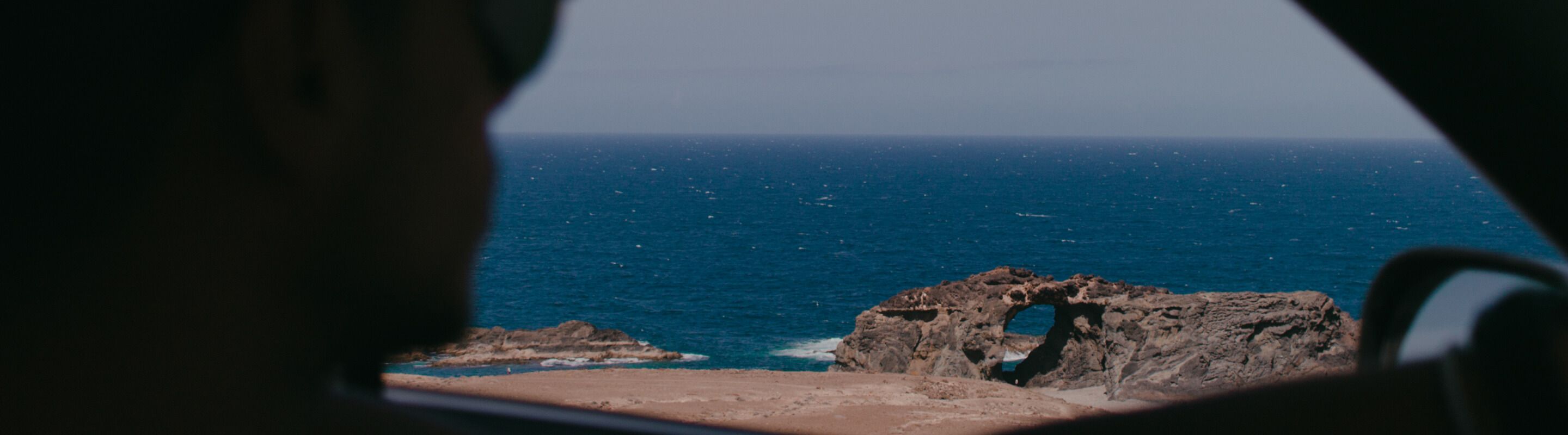 Foto do oceano visto do assento do passageiro de um carro, com o motorista embaçado.