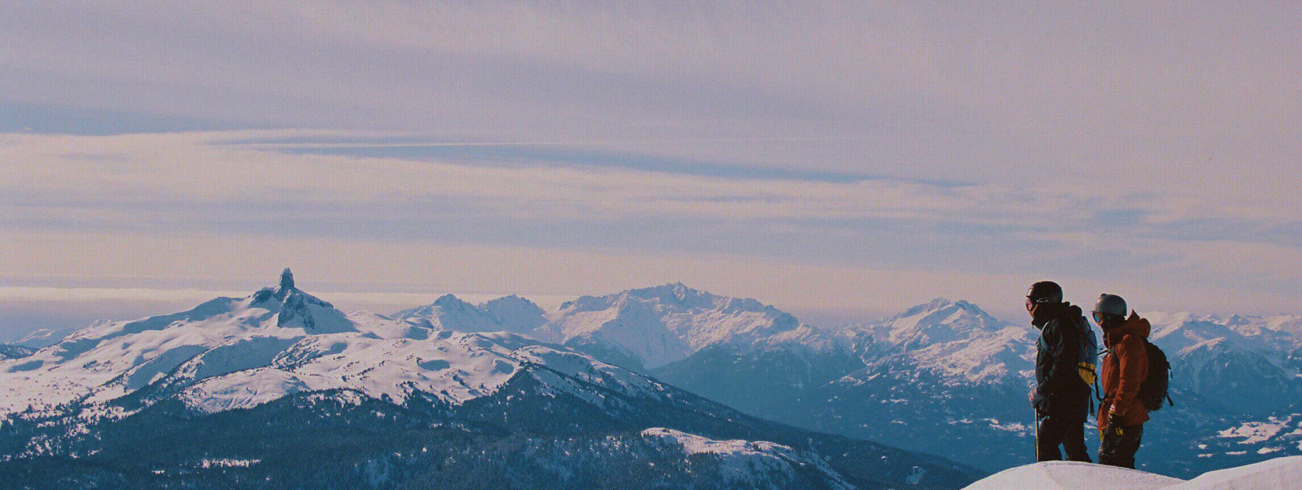 Two people stand on snow-covered mountain looking down at snow-capped mountains and forest below.